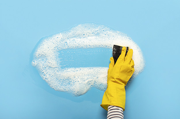 Hand in a yellow rubber glove holds a cleaning sponge and wipes a soapy foam on a blue background. Cleaning concept, cleaning service. Banner. Flat lay, top view